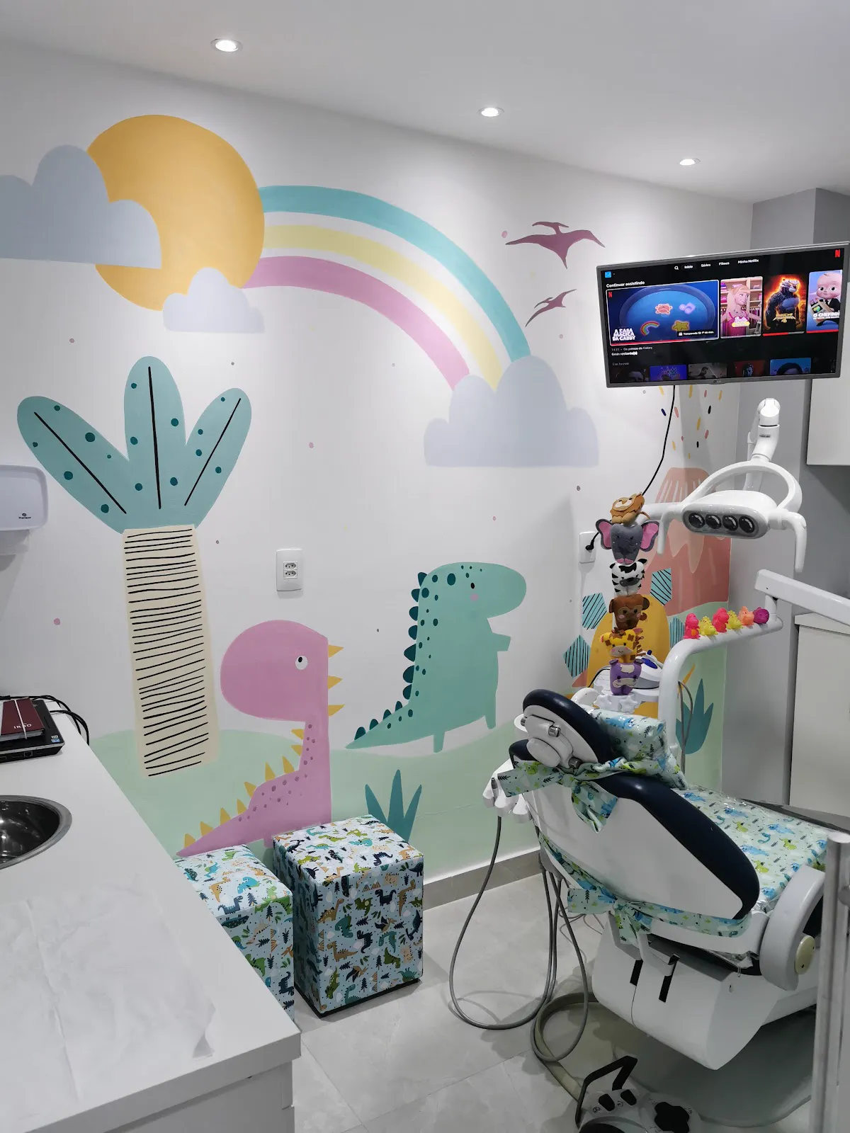 A cadeira do consultório diante da parede pintada com dinossauros e arco-íris, e a TV acesa no alto.